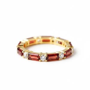 May include: A gold ring with alternating rectangular red and round, clear gemstones. The ring's design is set against a white background. The gemstones' colours are highlighted in this piece of jewellery.