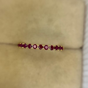 May include: A gold ring featuring a line of bright red gemstones. The ring is displayed against a neutral backdrop, accentuating the colour and sparkle of the gems. The ring is a delicate piece of jewellery.