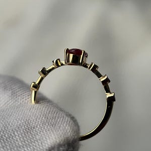 May include: A gold ring with a small, round, red gemstone at the center. The band features a series of small, rectangular accents. The ring is held by a hand wearing a white glove.