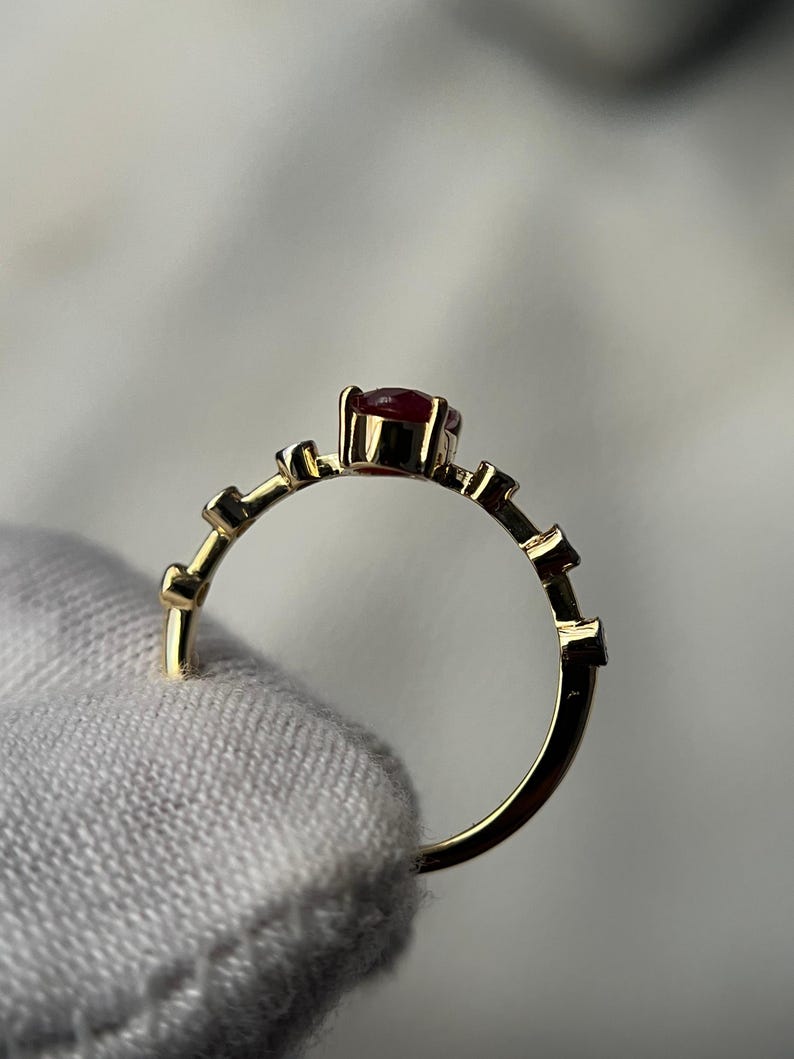 May include: A gold ring with a small, round, red gemstone at the center. The band features a series of small, rectangular accents. The ring is held by a hand wearing a white glove.