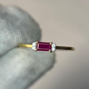 May include: A gold ring featuring a rectangular baguette-cut deep red ruby center stone flanked by two small round white diamonds on each side, set in a prong setting on a thin, polished gold band that creates a delicate and refined appearance.