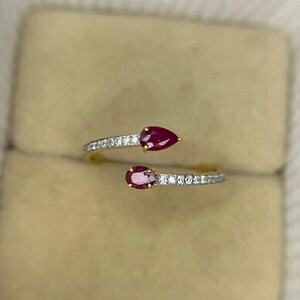 Op de afbeelding: Een goudkleurige ring met twee peervormige, dieprode edelstenen. De ringband is versierd met kleine, sprankelende witte stenen. Het ontwerp is open, met de edelstenen aan weerszijden van de band.