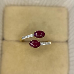 Puede incluir: Un anillo de oro con dos rubíes ovales de color rojo intenso. La banda se divide, cada lado adornado con pequeñas piedras blancas brillantes. El anillo se muestra sobre un fondo neutro, resaltando el diseño de la joya y el color de los rubíes.