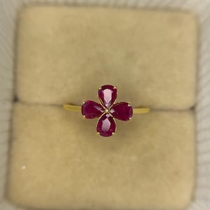 Könnte beinhalten: Ein goldener Ring mit einem vierblättrigen Blumenmuster. Jedes Blütenblatt ist ein leuchtender, birnenförmiger Rubin, der einen auffälligen Kontrast zum Goldband bildet. Der Ring ist vor einem neutralen, beigefarbenen Hintergrund platziert.