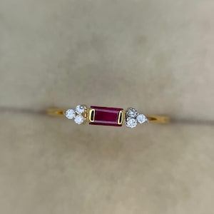 Puede incluir: Un anillo de oro con un rubí rojo central de corte baguette rectangular engastado en un bisel de oro. El rubí está flanqueado a ambos lados por un grupo de tres pequeños diamantes blancos redondos engastados en metal plateado, creando un diseño equilibrado.