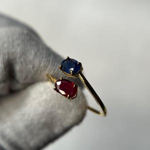 May include: A gold-colored ring featuring two gemstones: a deep blue sapphire and a vibrant red ruby. The gemstones are oval-shaped and set in a delicate, open design. The ring is shown against a neutral background.