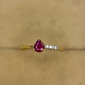 May include: A gold ring with a pear-cut, deep red gemstone. The band splits, with small, clear stones set on one side. The ring is set against a neutral background, highlighting the jewelry's design. A classic piece of jewellery.