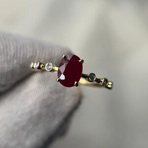 May include: A gold ring featuring a large, oval-cut ruby gemstone as the centerpiece. The ring is accented with small, round, clear gemstones. The ring's band has a delicate design, adding to its elegant appearance.