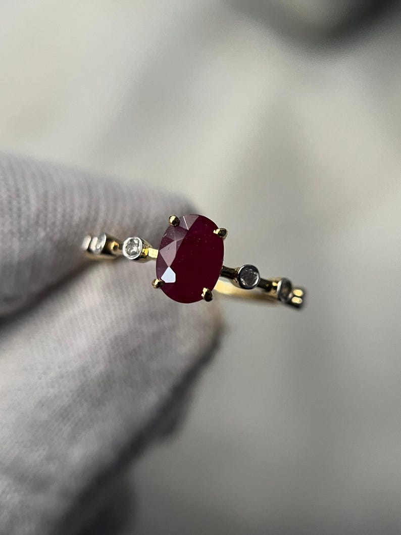 May include: A gold ring featuring a large, oval-cut ruby gemstone as the centerpiece. The ring is accented with small, round, clear gemstones. The ring's band has a delicate design, adding to its elegant appearance.