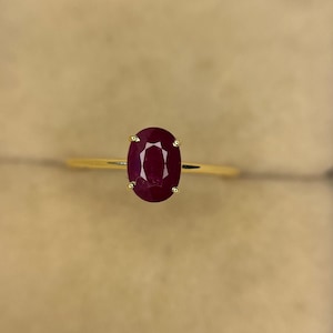 Könnte beinhalten: Ein goldener Ring mit einem ovalen, tiefroten Rubin-Edelstein. Der Rubin wird von vier Krappen gehalten, und das Band ist aus dünnem, poliertem Gold. Der Ring wird vor einem neutralen Hintergrund gezeigt.