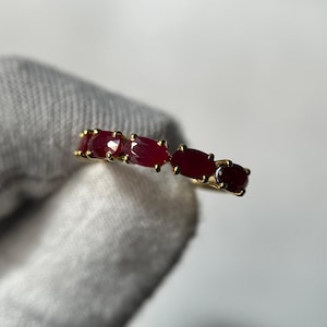May include: A gold ring featuring five oval-cut, deep red gemstones. The gemstones are set in a row along the band. The ring is held against a white background, with a soft, neutral-colored fabric in the foreground.