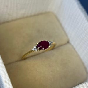 May include: A gold ring with an oval ruby gemstone and small diamond accents. The ring is set in a delicate gold band and is presented in a cream-coloured ring box.