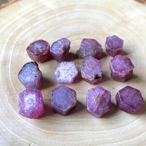 May include: A collection of raw ruby gemstones in various shapes and sizes. The gemstones display a range of pink and purple hues, with some showing darker red tones. They are arranged on a light brown, wooden surface.