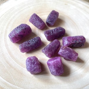 Puede incluir: Una colección de diez piedras preciosas de rubí en bruto de color púrpura, dispuestas sobre una superficie de madera. Las piedras varían en tamaño y forma, algunas son más puntiagudas y otras más redondeadas.