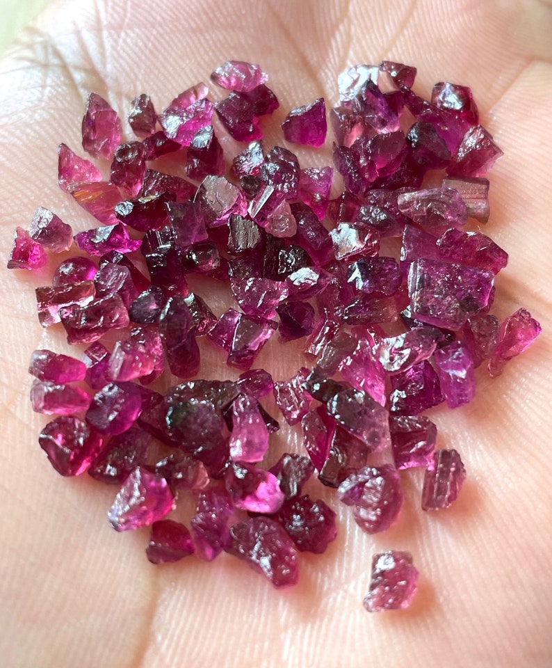 May include: A collection of loose pink and purple gemstones, likely rubies or sapphires, in various shapes and sizes. The gemstones are scattered on a hand, showcasing their natural beauty and brilliance.