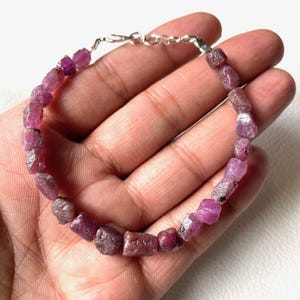 Könnte beinhalten: Ein Armband aus ungeschliffenen, rosaroten Edelsteinen. Die Steine sind mit einer silberfarbenen Kette und einem Verschluss verbunden. Das Armband wird vor einem hellen Hintergrund präsentiert.