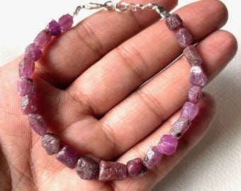 Op de afbeelding: Een armband gemaakt van ruwe, rozerode edelstenen. De stenen zijn geregen met een zilverkleurige ketting en sluiting. De armband wordt gepresenteerd tegen een lichte achtergrond.