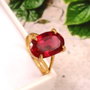 Könnte beinhalten: Ein goldener Ring mit einem großen, ovalen, roten Edelstein. Der Ring hat ein schlichtes, elegantes Design.