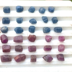 May include: A collection of 30 loose gemstones, including blue sapphires and red rubies, arranged in rows on a white surface.