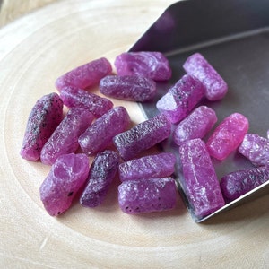 Peut inclure: Une collection de pierres précieuses de rubis brutes de couleur violette, certaines avec des inclusions noires, éparpillées sur une surface en bois. Les pierres précieuses sont de tailles et de formes variées, avec quelques-unes dans une petite pelle en métal.