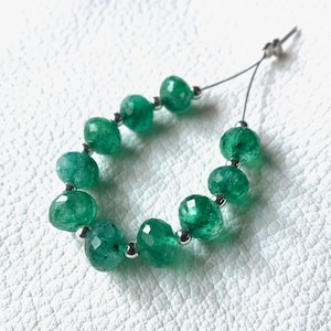 Op de afbeelding: Een streng gefacetteerde smaragdgroene kralen, elk met een levendige groene tint, geregen met een zilverkleurige draad. De kralen zijn rond en hebben een gepolijst, reflecterend oppervlak. De achtergrond is een wit oppervlak met textuur.