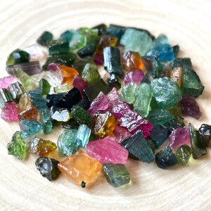 May include: A collection of raw, rough, and tumbled gemstones in various colors, including green, blue, pink, and brown. The gemstones are scattered on a wooden surface.