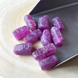 May include: A group of ten rough, pink, purple, and red gemstones. The gemstones are in various shapes and sizes and are arranged on a wooden surface.