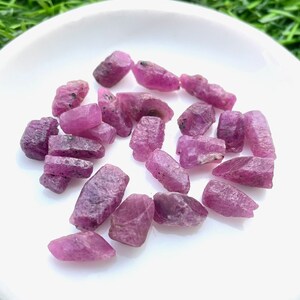 Pu&ograve; includere: Una collezione di pietre grezze di rubino rosa su un piatto bianco. Le pietre sono di varie dimensioni e forme, alcune hanno una forma pi&ugrave; appuntita e altre sono pi&ugrave; arrotondate.