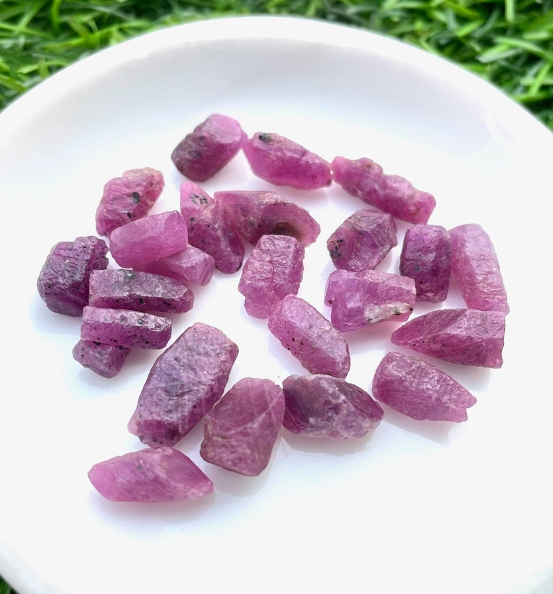 Pu&ograve; includere: Una collezione di pietre grezze di rubino rosa su un piatto bianco. Le pietre sono di varie dimensioni e forme, alcune hanno una forma pi&ugrave; appuntita e altre sono pi&ugrave; arrotondate.