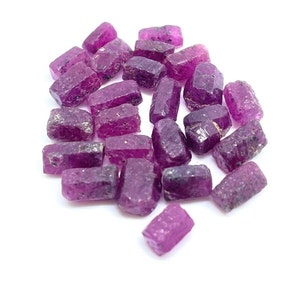 Könnte beinhalten: Eine Ansammlung roher, ungeschliffener Rubin-Edelsteine. Die Steine haben eine tiefe, satte magenta Farbe und raue, strukturierte Oberflächen. Sie liegen auf einem hellen, weißen Hintergrund, was ihre natürliche Schönheit und ihr Potenzial für Schmuck oder Sammlungen hervorhebt.