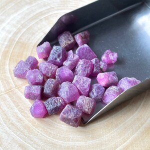 May include: A collection of raw, uncut ruby gemstones in varying sizes and shapes. The rubies display a vibrant pinkish-red hue, with some showing darker, purplish tones. They are presented in a small, silver scoop, resting on a light-colored wooden surface.