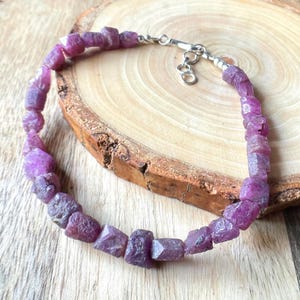 May include: A silver chain bracelet with faceted purple ruby beads. The bracelet is on a wooden surface.