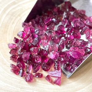 May include: A close-up of a small pile of pink and purple gemstone chips. The chips are scattered on a wooden surface and are being poured from a metal scoop.