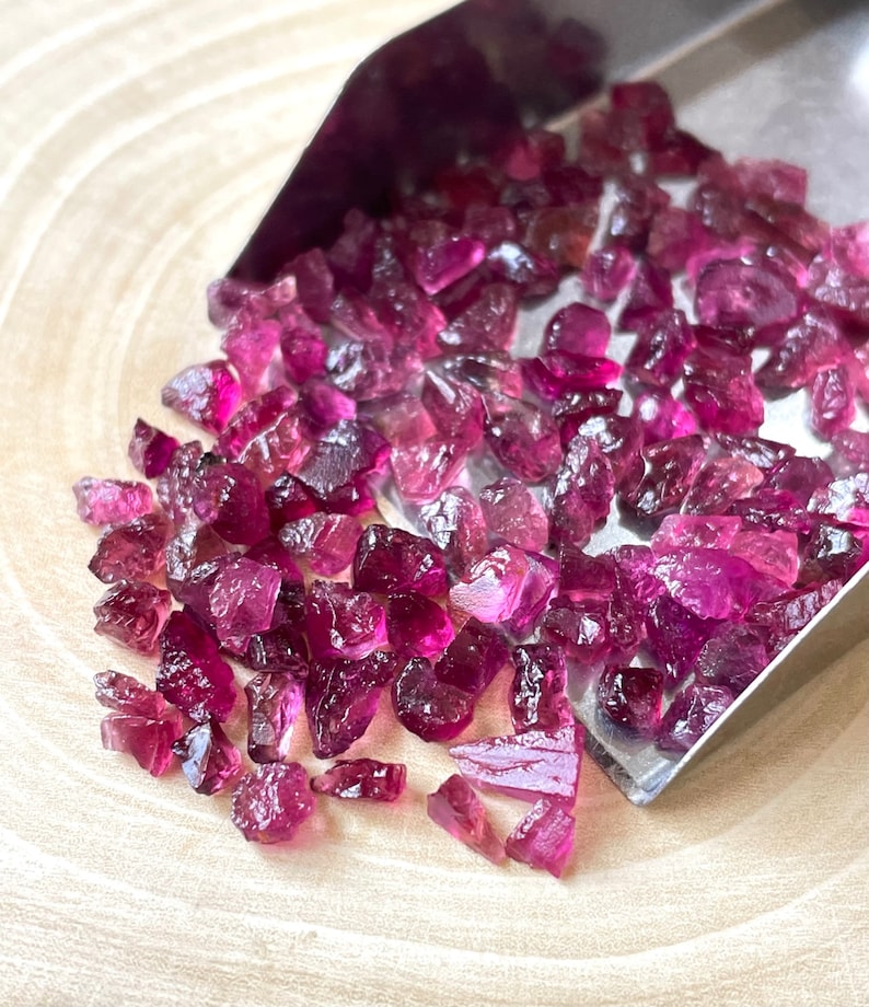 May include: A close-up of a small pile of pink and purple gemstone chips. The chips are scattered on a wooden surface and are being poured from a metal scoop.