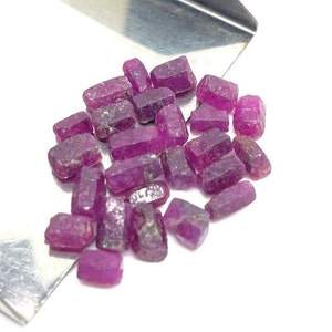 May include: A collection of raw, uncut ruby gemstones. The rubies are a deep pinkish-red color, with a rough, textured surface. They are scattered on a white surface, with a silver scoop in the background.