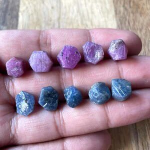 May include: A collection of eight faceted gemstones, four pink and four blue, arranged in two rows on a hand. The gemstones are likely rubies and sapphires.