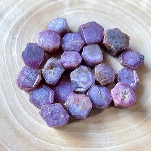 May include: A cluster of raw ruby crystals, displaying a rich purplish-red hue. The gemstones have a hexagonal form with visible facets. They are presented on a light-toned wooden surface, highlighting their natural appearance.