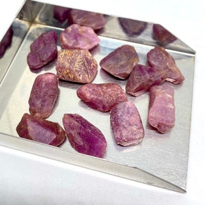 Puede incluir: Una colección de piedras brutas de rubí rosa en una bandeja de metal. Las piedras brutas son de varios tamaños y formas.