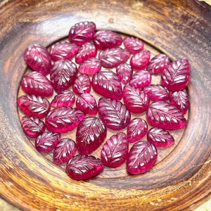 Puede incluir: Una colección de piedras preciosas de color rojo rubí en forma de hoja en un cuenco de madera. Las piedras preciosas están facetadas y tienen un acabado pulido.