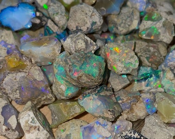 Puede incluir: Una colección de piedras de ópalo en bruto y sin pulir en varios tonos de gris, azul, verde y blanco. Las piedras tienen un brillo natural e iridiscente.