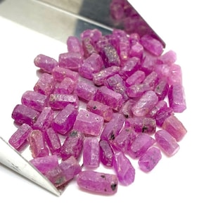 Könnte beinhalten: Eine Sammlung von rosafarbenen Rubingranaten, jede mit einer rechteckigen Form und einer rauen, natürlichen Oberfläche. Die Rubine sind auf einer silbernen Oberfläche verstreut.