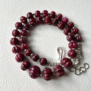 Op de afbeelding: Een ketting met dieprode, gesneden edelsteen kralen. De kralen zijn geregen met kleine zilverkleurige kralen en een zilverkleurige sluiting. De ketting wordt gepresenteerd op een wit oppervlak.