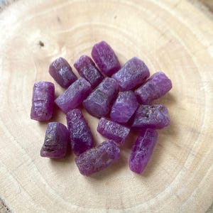 Puede incluir: Un conjunto de gemas de rubí en bruto sin cortar. Las piedras son de un color magenta intenso y rico, con una superficie rugosa y texturizada. Están dispuestas sobre una superficie de madera marrón claro.