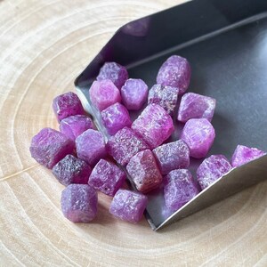 Op de afbeelding: Een verzameling ruwe, ongeslepen robijn edelstenen in verschillende tinten roze en paars. De edelstenen worden gepresenteerd op een houten oppervlak, met een gedeeltelijk zichtbare metalen schep. De ruwe textuur van de stenen is duidelijk zichtbaar.