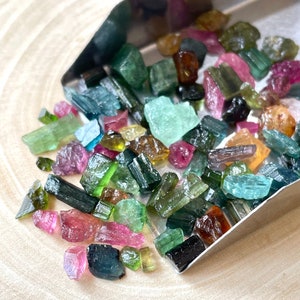 May include: A collection of colorful, raw, rough, and tumbled gemstones. The gemstones are various shades of green, blue, pink, and brown. The gemstones are in a metal scoop on a wooden surface.