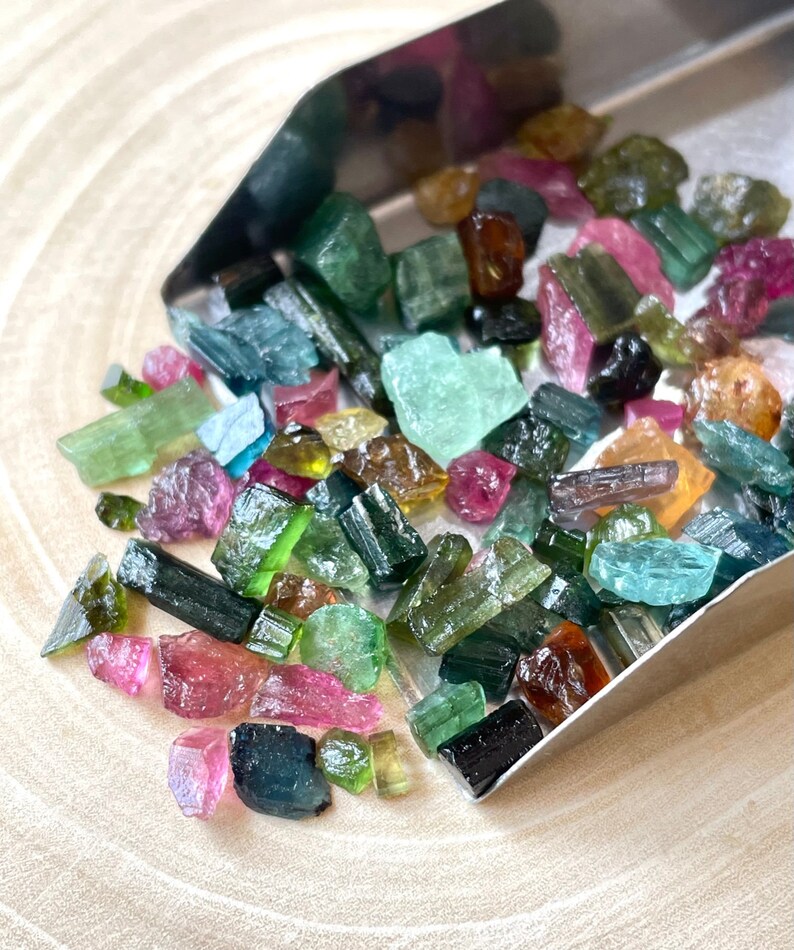 May include: A collection of colorful, raw, rough, and tumbled gemstones. The gemstones are various shades of green, blue, pink, and brown. The gemstones are in a metal scoop on a wooden surface.