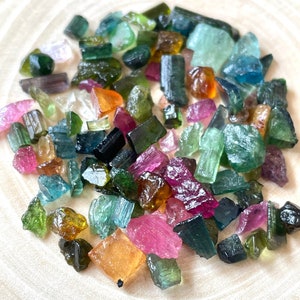 May include: A collection of raw, rough, and tumbled gemstones in various colors, including green, blue, pink, and brown. The gemstones are scattered on a light wood surface.
