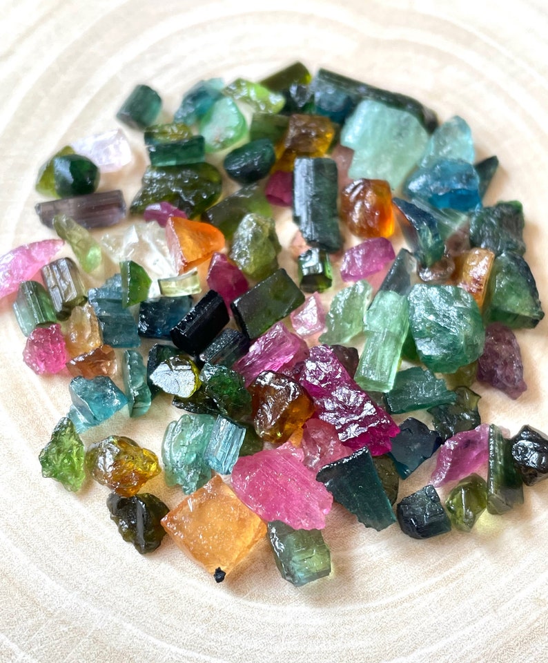 May include: A collection of raw, rough, and tumbled gemstones in various colors, including green, blue, pink, and brown. The gemstones are scattered on a light wood surface.