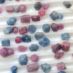 Peut inclure: Une collection de pierres précieuses facettées en vrac dans des tons de bleu et de rose. Les pierres précieuses sont disposées sur une surface blanche.