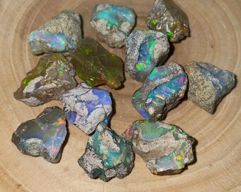 Puede incluir: Una colección de piedras de ópalo en bruto con colores iridiscentes, incluyendo verde, azul y morado. Las piedras son de varios tamaños y formas, y algunas tienen una matriz gris.